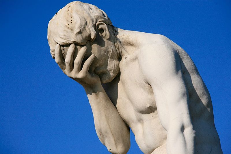 Close-up of a white marble statue of a man covering his face with his hand, set against a bright blue sky, conveying frustration